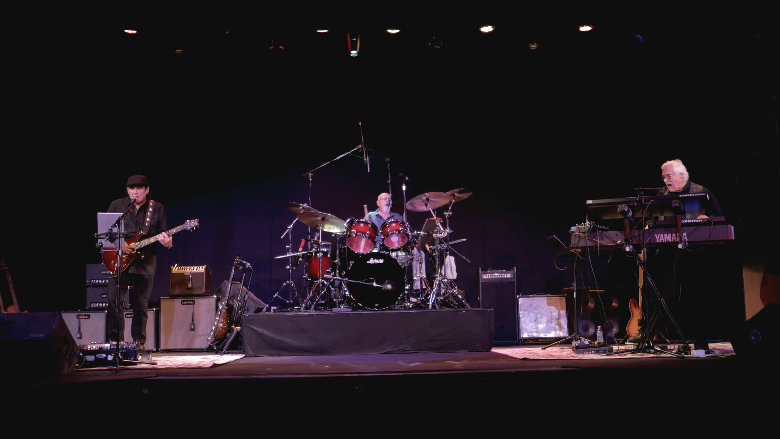 Allen playing a red guitar at left, with Ed on drums and Rico at the keyboards on the right.
